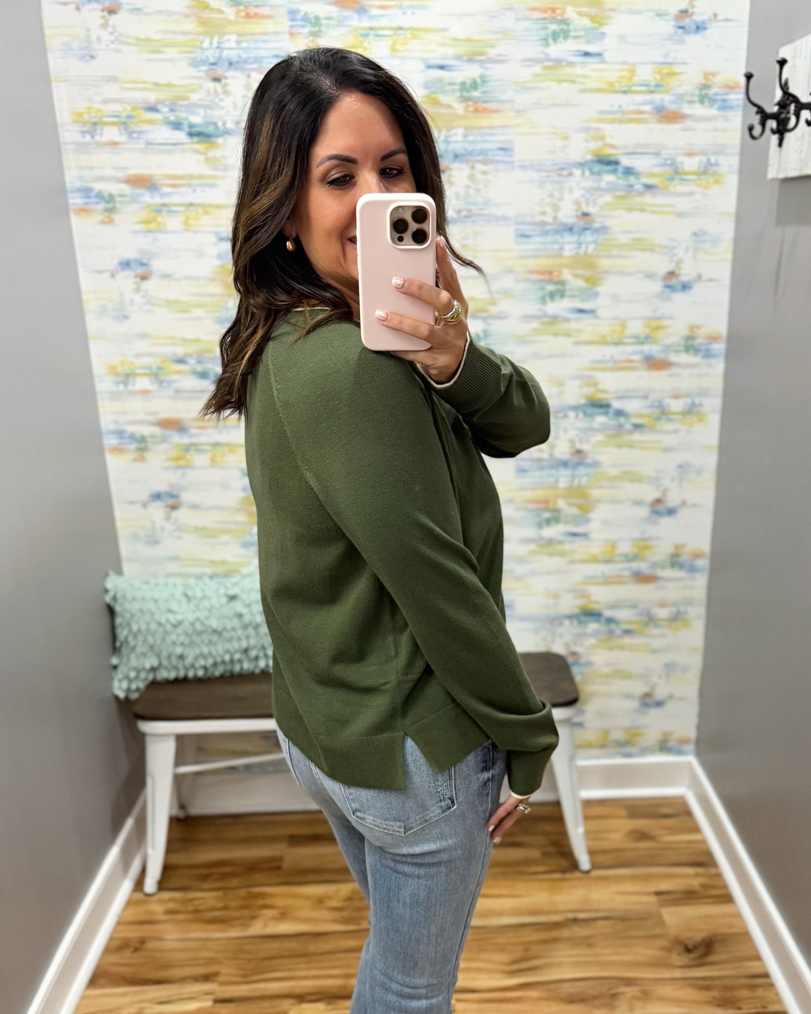 A woman takes a mirror selfie wearing an olive green long-sleeve knit top and light blue jeans in a room with patterned wallpaper and a wooden floor.