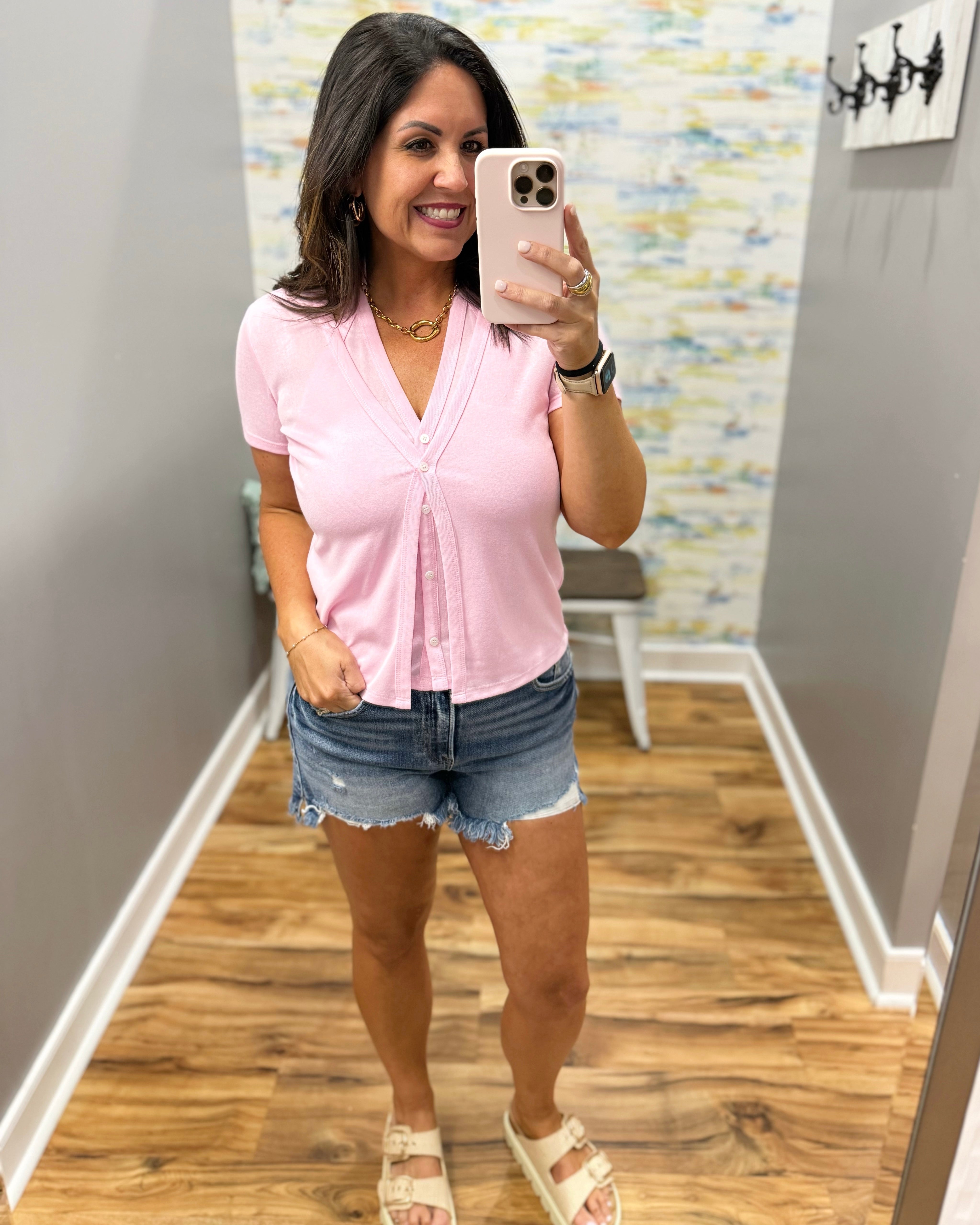 Woman taking a mirror selfie wearing a pink top and denim shorts in a room with wooden flooring and a patterned wall.