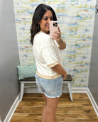 Woman taking a mirror selfie wearing a white top and denim shorts in a room with a textured wall.