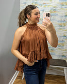 A woman takes a mirror selfie wearing a brown halter-neck ruffled top and dark blue jeans in a fitting room.