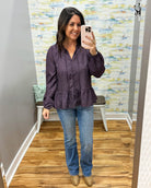 A woman takes a mirror selfie wearing a dark purple patterned long-sleeve blouse with a peplum hem, paired with blue jeans and tan boots, in a fitting room with a wood floor and patterned wallpaper.