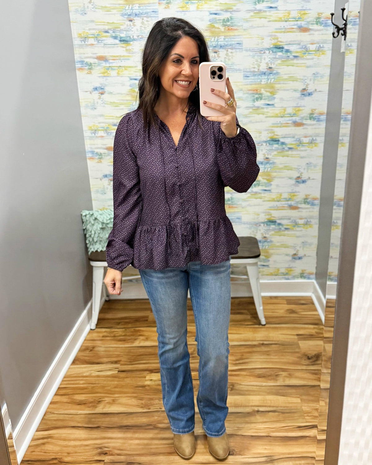 A woman takes a mirror selfie wearing a dark purple patterned long-sleeve blouse with a peplum hem, paired with blue jeans and tan boots, in a fitting room with a wood floor and patterned wallpaper.