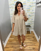 A woman takes a mirror selfie in a fitting room wearing a beige and black vertically striped short-sleeve mini dress with a V-neckline.