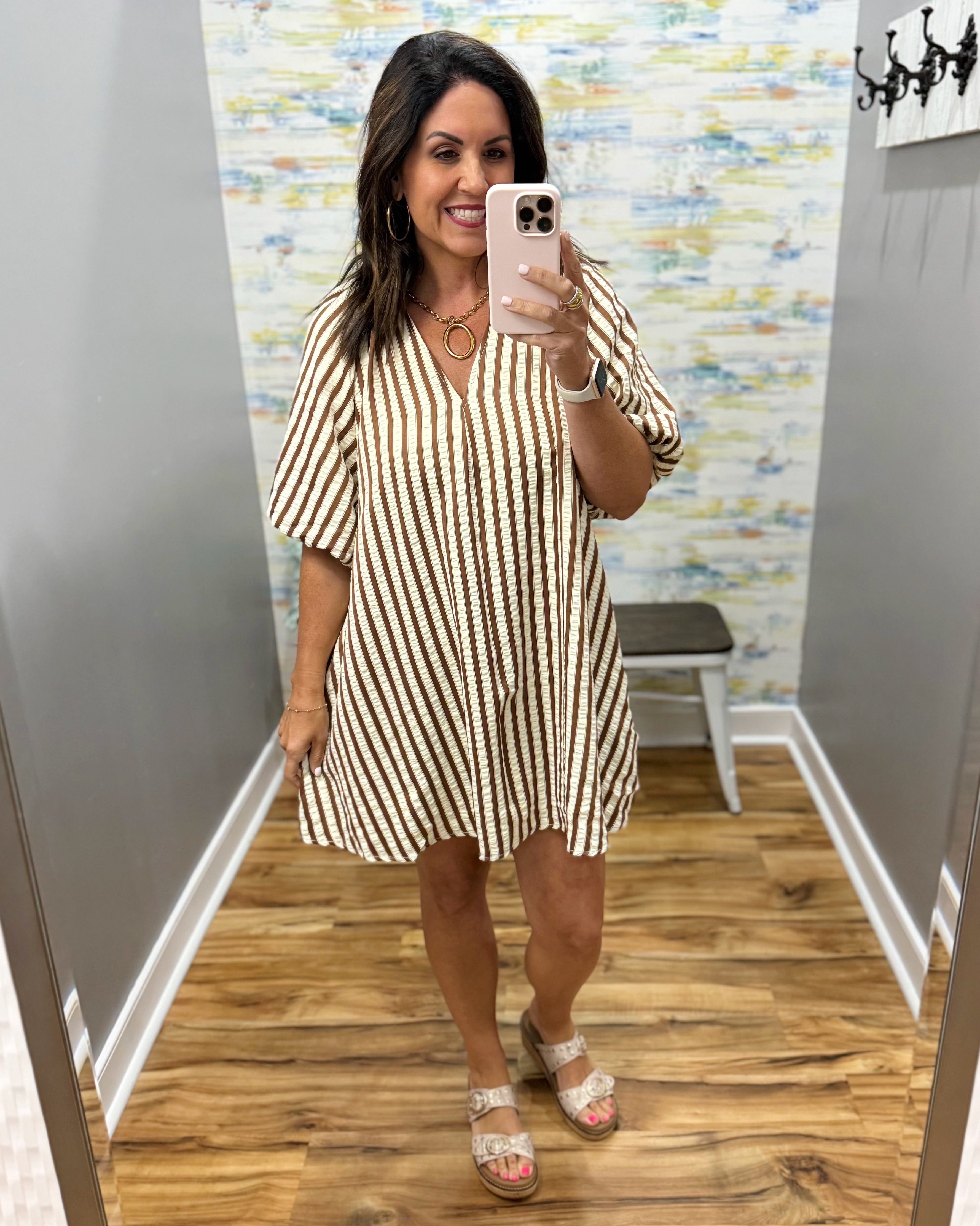 A woman takes a mirror selfie in a fitting room wearing a beige and black vertically striped short-sleeve mini dress with a V-neckline.