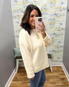 A woman takes a mirror selfie wearing a cream-colored knit sweater and blue jeans in a home setting with a patterned wallpaper background.