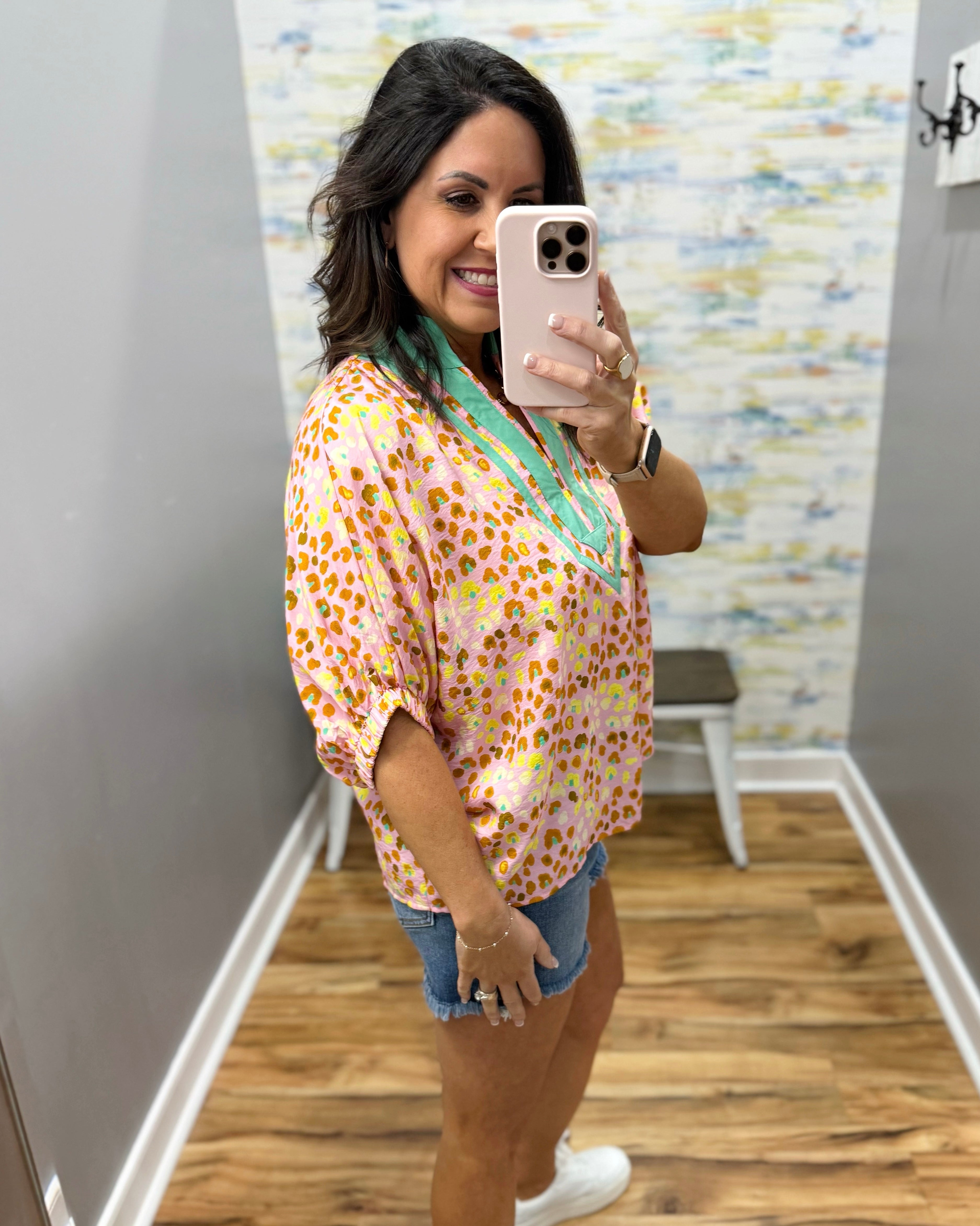 Woman taking a mirror selfie wearing a floral blouse and denim shorts in a room with a patterned wall.