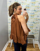 A woman takes a mirror selfie wearing a sleeveless rust-brown ruffled top with a tie detail at the back, paired with dark blue jeans.