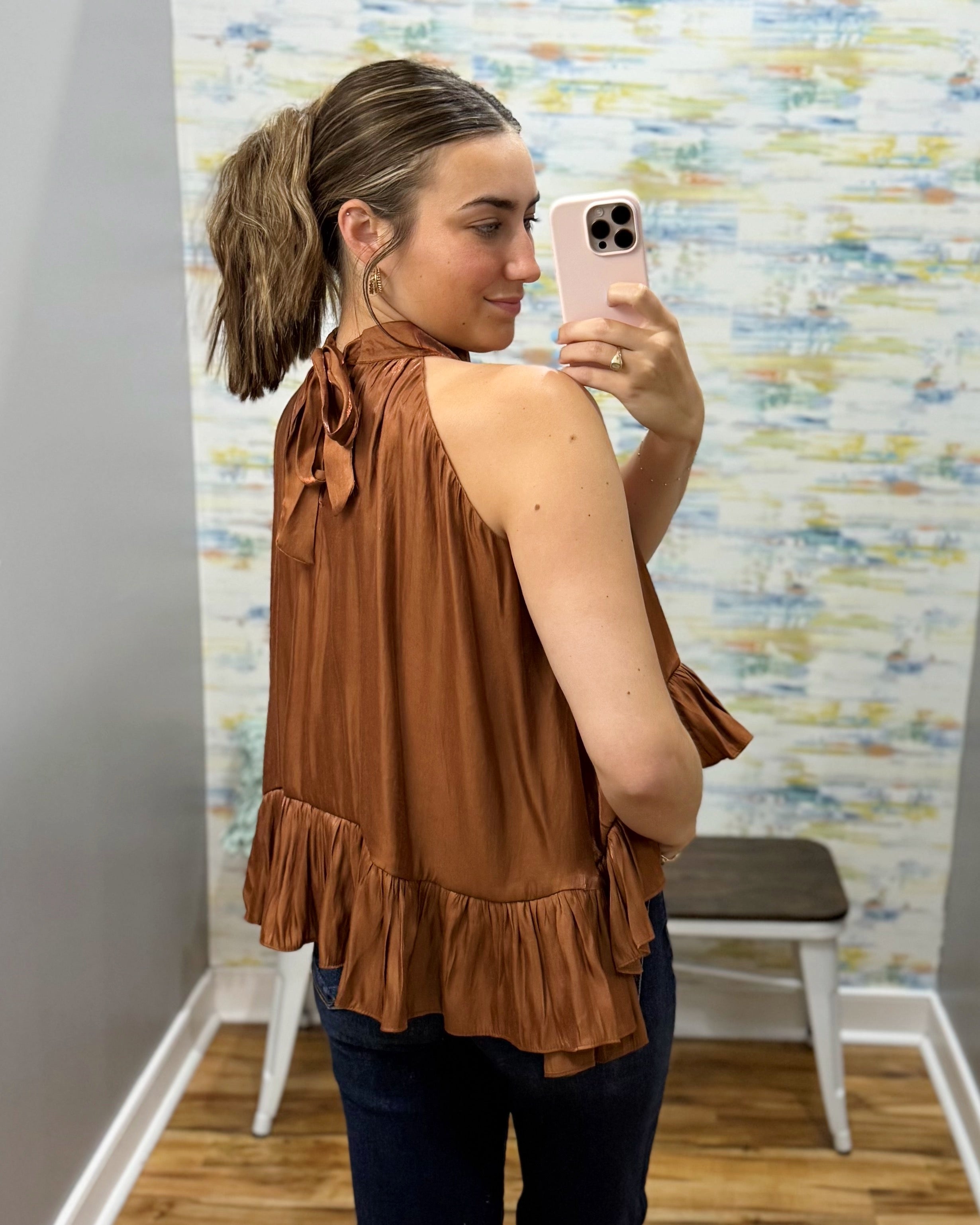 A woman takes a mirror selfie wearing a sleeveless rust-brown ruffled top with a tie detail at the back, paired with dark blue jeans.