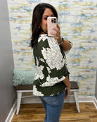 A woman takes a mirror selfie in a fitting room wearing a dark green blouse with a large white floral pattern and blue jeans.