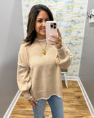 A woman takes a mirror selfie wearing a beige knit sweater with white contrast trim on the collar, cuffs, and hem, paired with blue jeans.