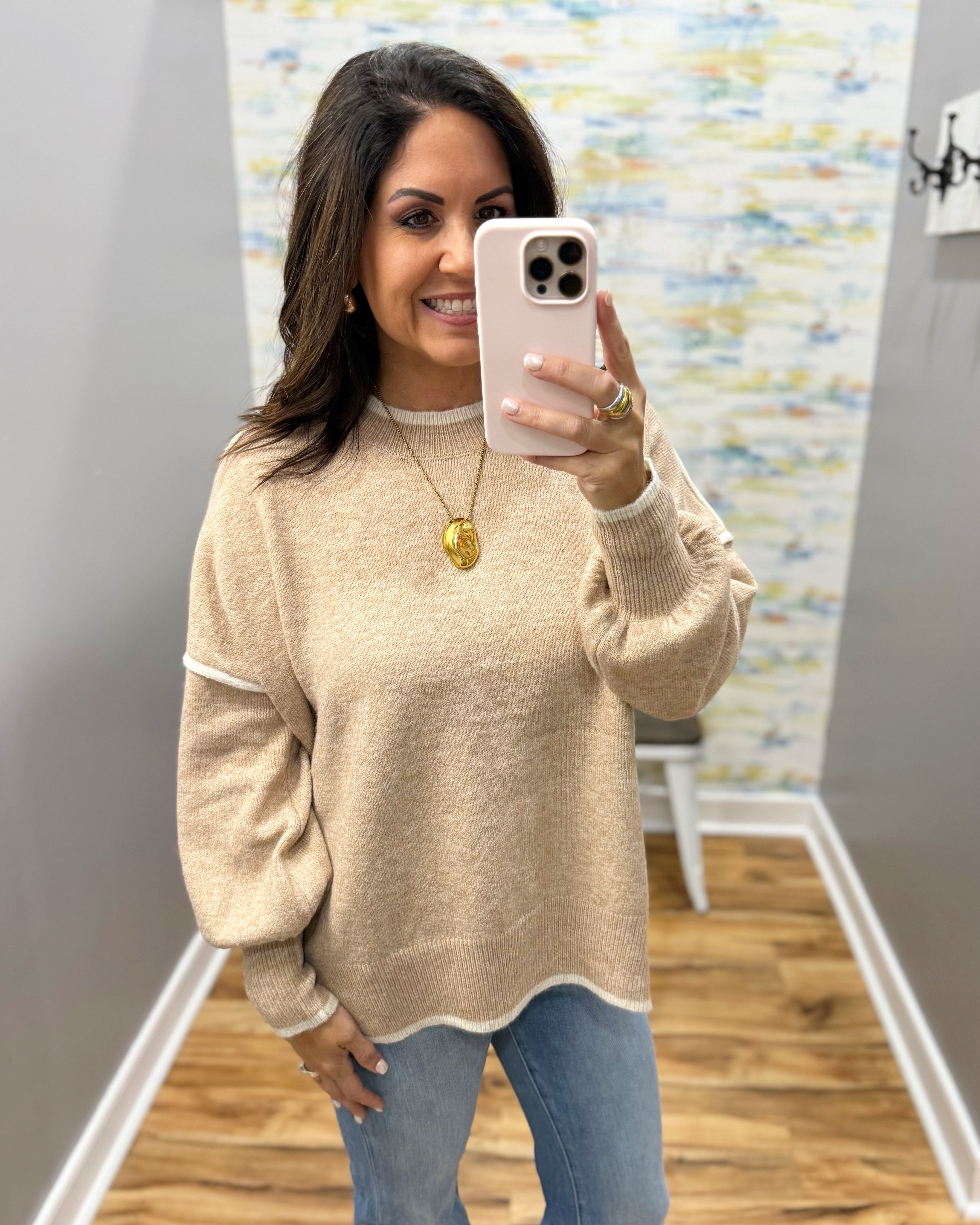 A woman takes a mirror selfie wearing a beige knit sweater with white contrast trim on the collar, cuffs, and hem, paired with blue jeans.