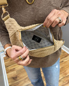 A person wearing a brown knit sweater and blue jeans holds open a woven beige crossbody bag with gold-tone hardware, showing the interior lining and a black brand patch.