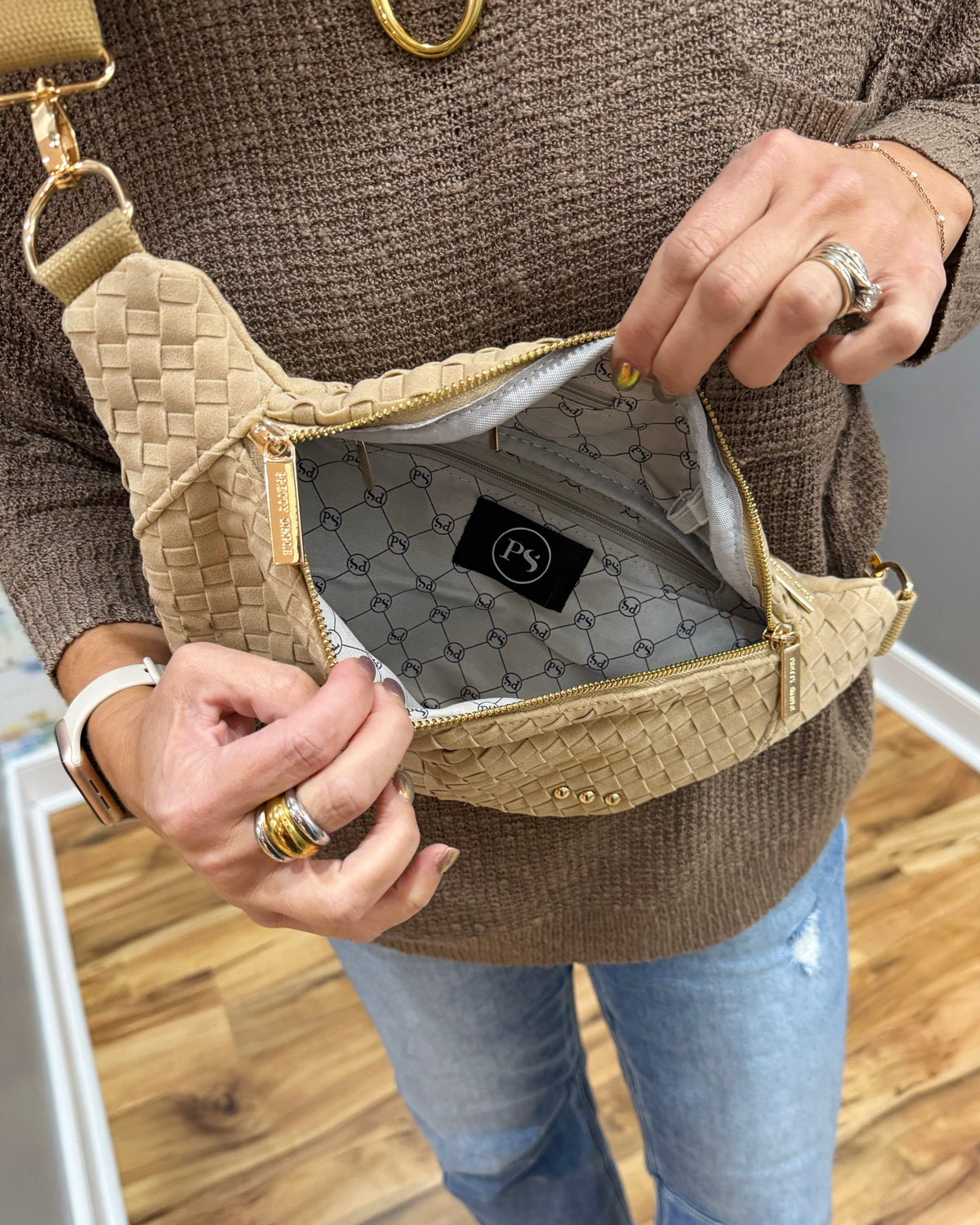 A person wearing a brown knit sweater and blue jeans holds open a woven beige crossbody bag with gold-tone hardware, showing the interior lining and a black brand patch.