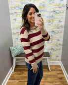 A woman takes a mirror selfie wearing a beige and burgundy striped knit sweater with blue jeans, standing in a room with a patterned wallpaper background.