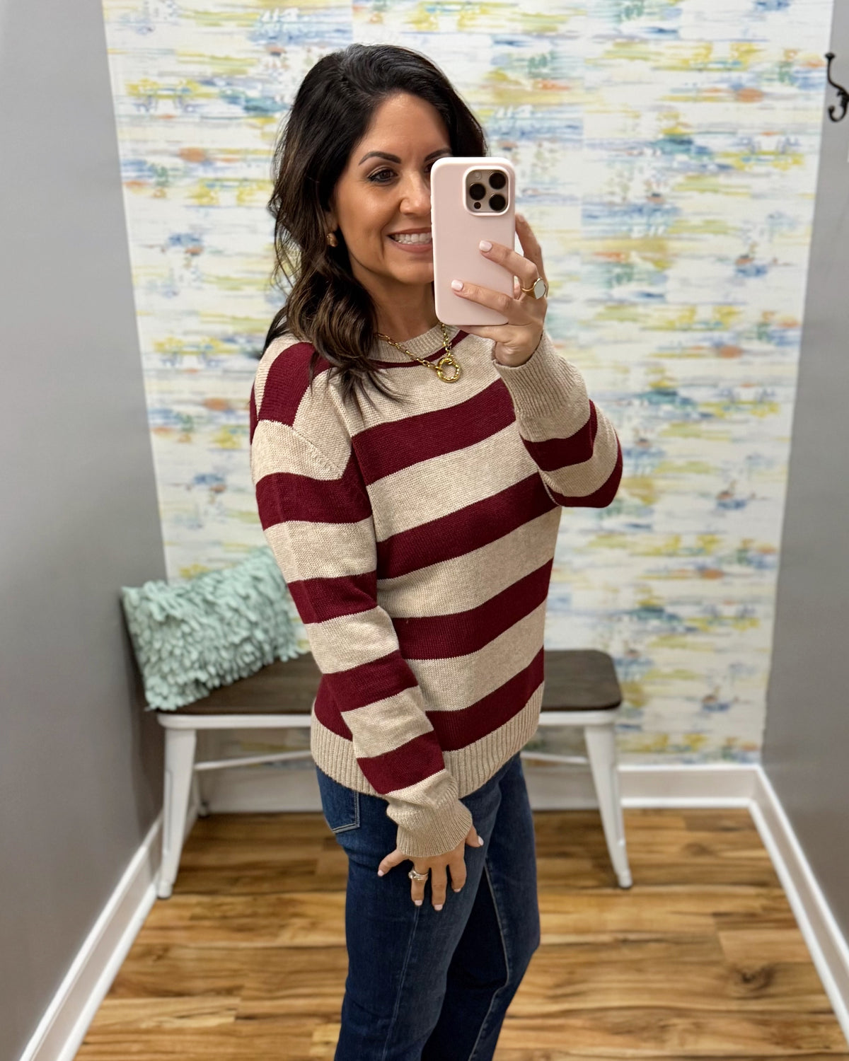 A woman takes a mirror selfie wearing a beige and burgundy striped knit sweater with blue jeans, standing in a room with a patterned wallpaper background.