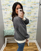 A woman takes a mirror selfie wearing a black and white striped knit sweater with a cream ribbed hem and cuffs, paired with blue jeans. The background features a colorful abstract wallpaper, a small bench with a mint green pillow, and a wood floor.