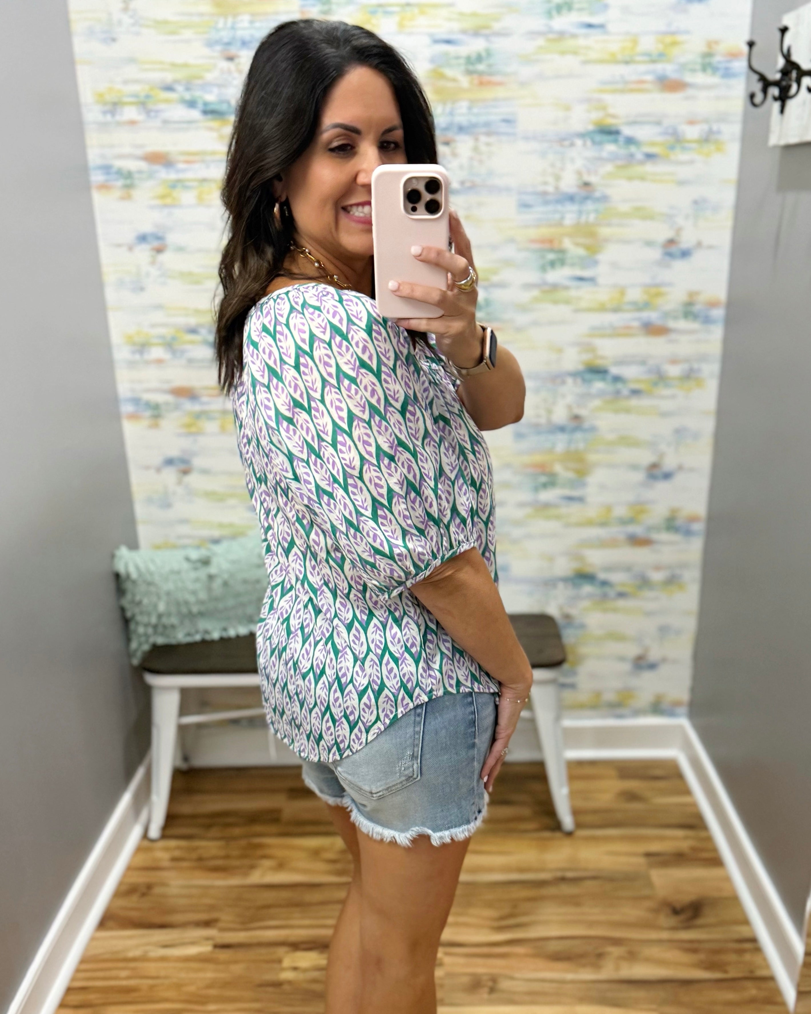 Woman taking a mirror selfie wearing a patterned top and denim shorts in a room with a textured wall.