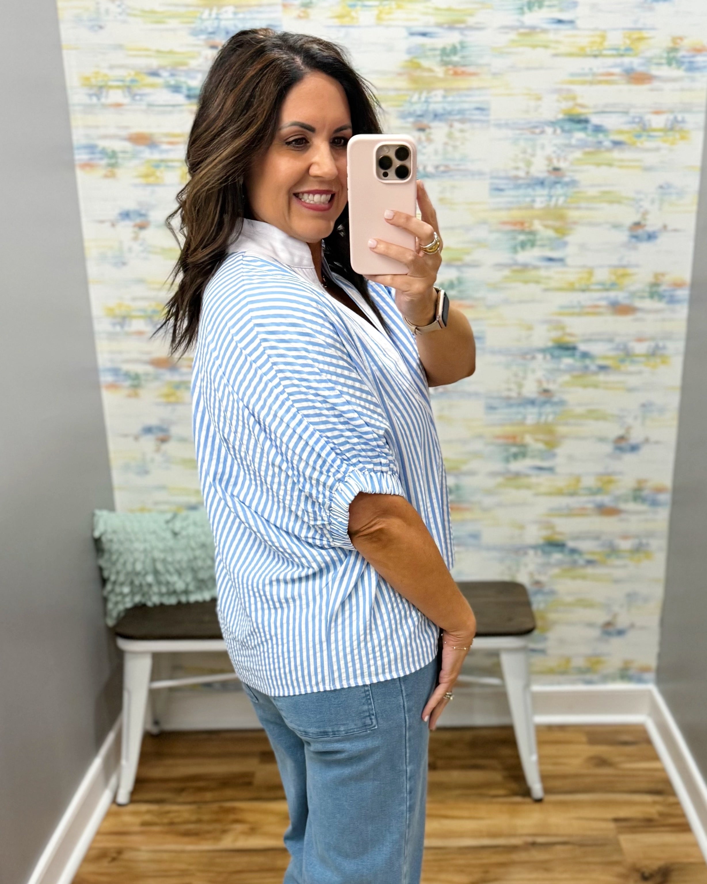 Woman taking a mirror selfie wearing a blue and white striped shirt and jeans, with a colorful wall in the background.
