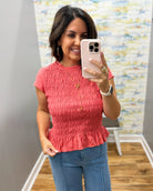 A woman takes a mirror selfie wearing a coral pink short-sleeve smocked peplum top and blue jeans in a fitting room.