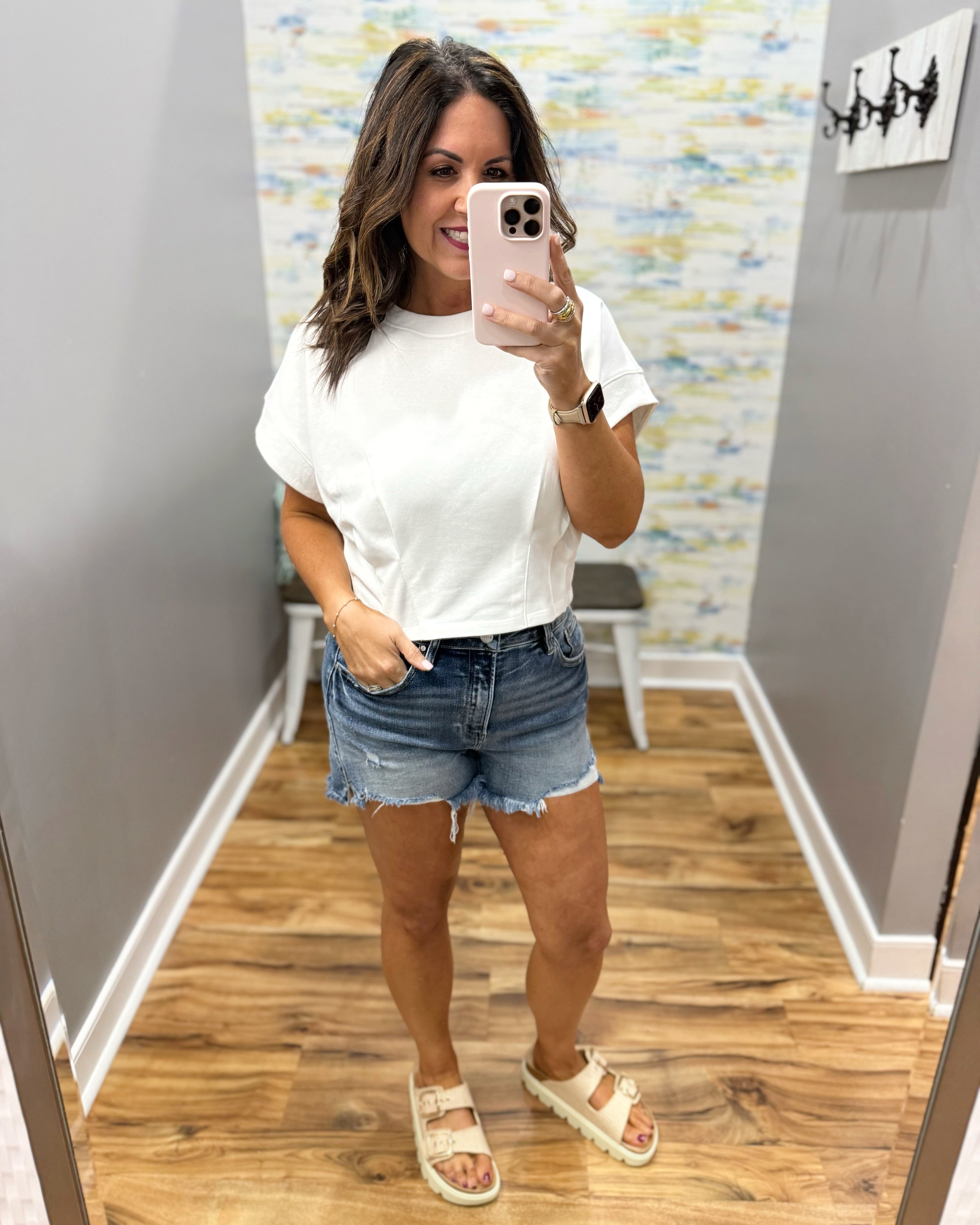 Woman taking a mirror selfie wearing a white t-shirt and denim shorts in a room with a colorful wall.