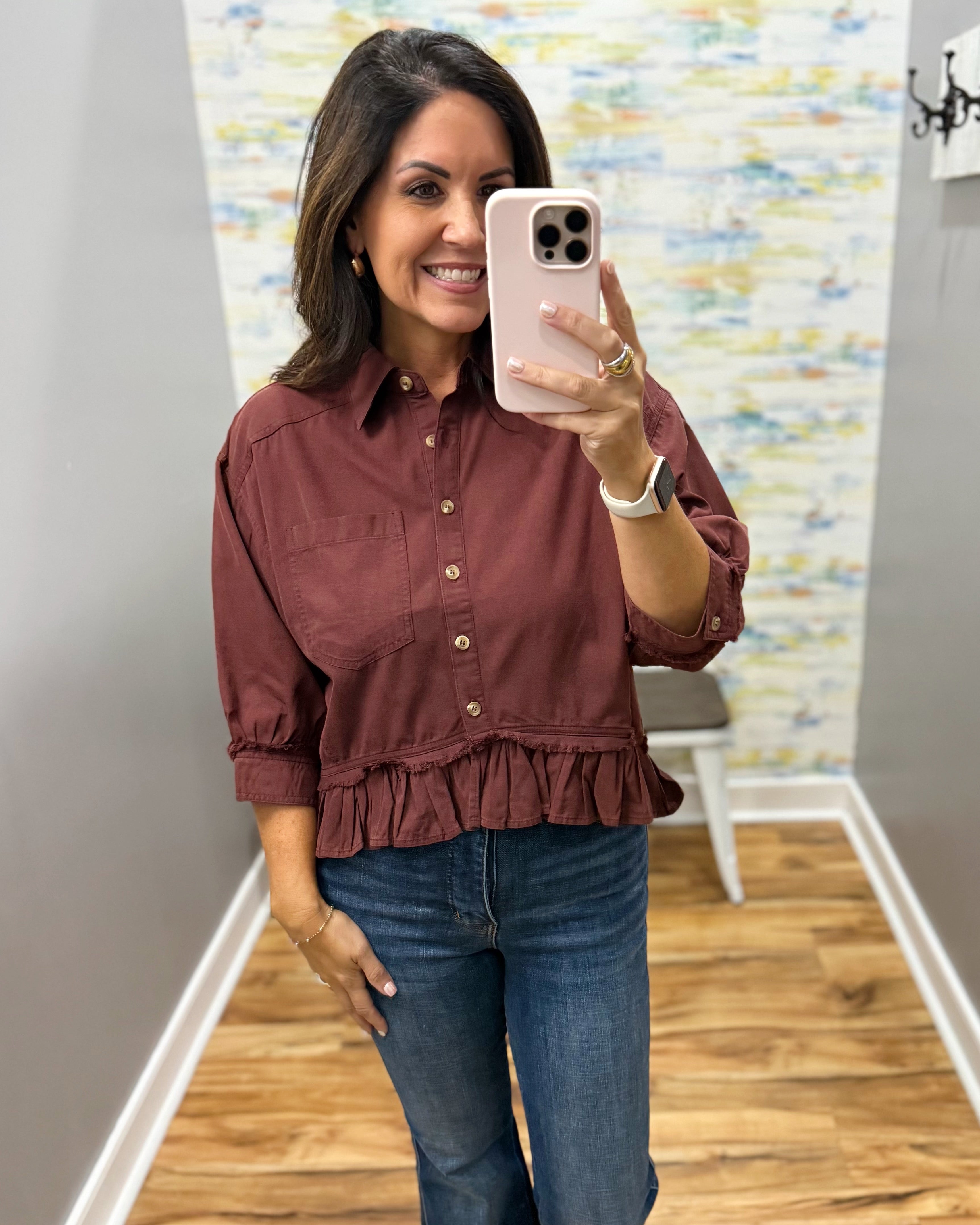 A woman takes a mirror selfie in an indoor hallway, wearing a rust-brown ruffled button-up shirt and blue jeans.