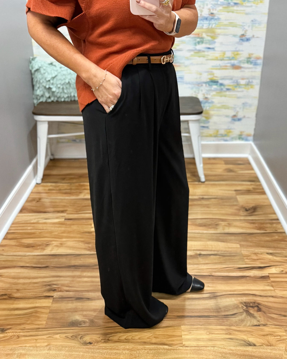 A person standing indoors wearing black wide-leg pants with a brown belt, paired with an orange short-sleeve top and black shoes. The photo is taken in a room with wood flooring, a bench, and a colorful wall mural.