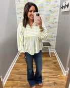 A woman takes a mirror selfie in a fitting room wearing a cream and green vertically striped long-sleeve blouse with a peplum waist, paired with dark blue flare jeans.