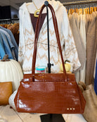 A brown crocodile-embossed leather handbag with double shoulder straps, displayed on a black stand in a clothing boutique setting.