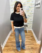 A woman takes a mirror selfie in an indoor hallway, wearing a black short-sleeve top with white ribbed trim at the collar and hem, paired with light blue flare jeans and beige flats.