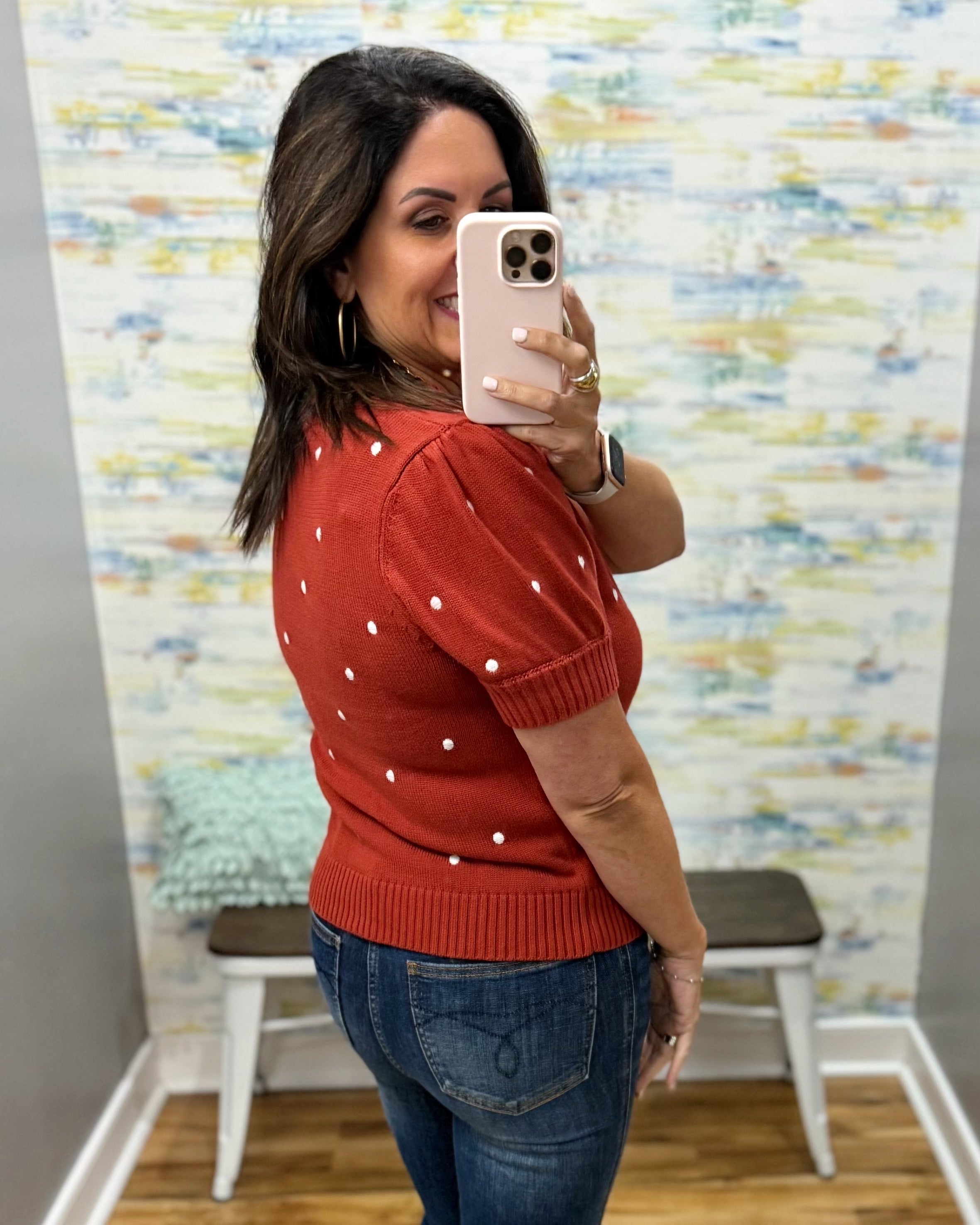 A woman takes a mirror selfie in a fitting room wearing a rust-red short-sleeve knit top with white polka dots and blue jeans.