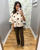 A woman takes a mirror selfie in a fitting room wearing a cream knit sweater with large black polka dots and dark brown wide-leg pants.
