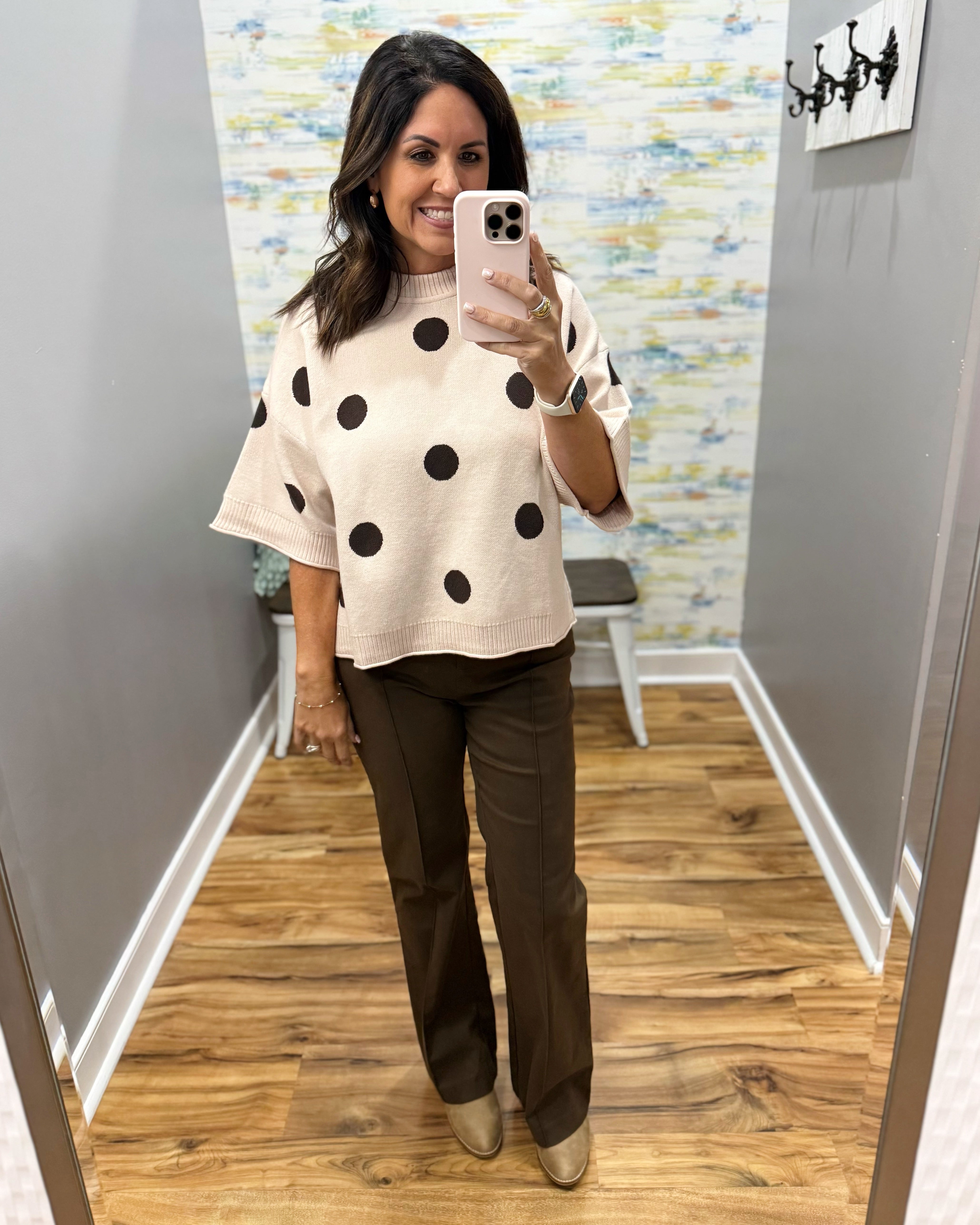 A woman takes a mirror selfie in a fitting room wearing a cream knit sweater with large black polka dots and dark brown wide-leg pants.