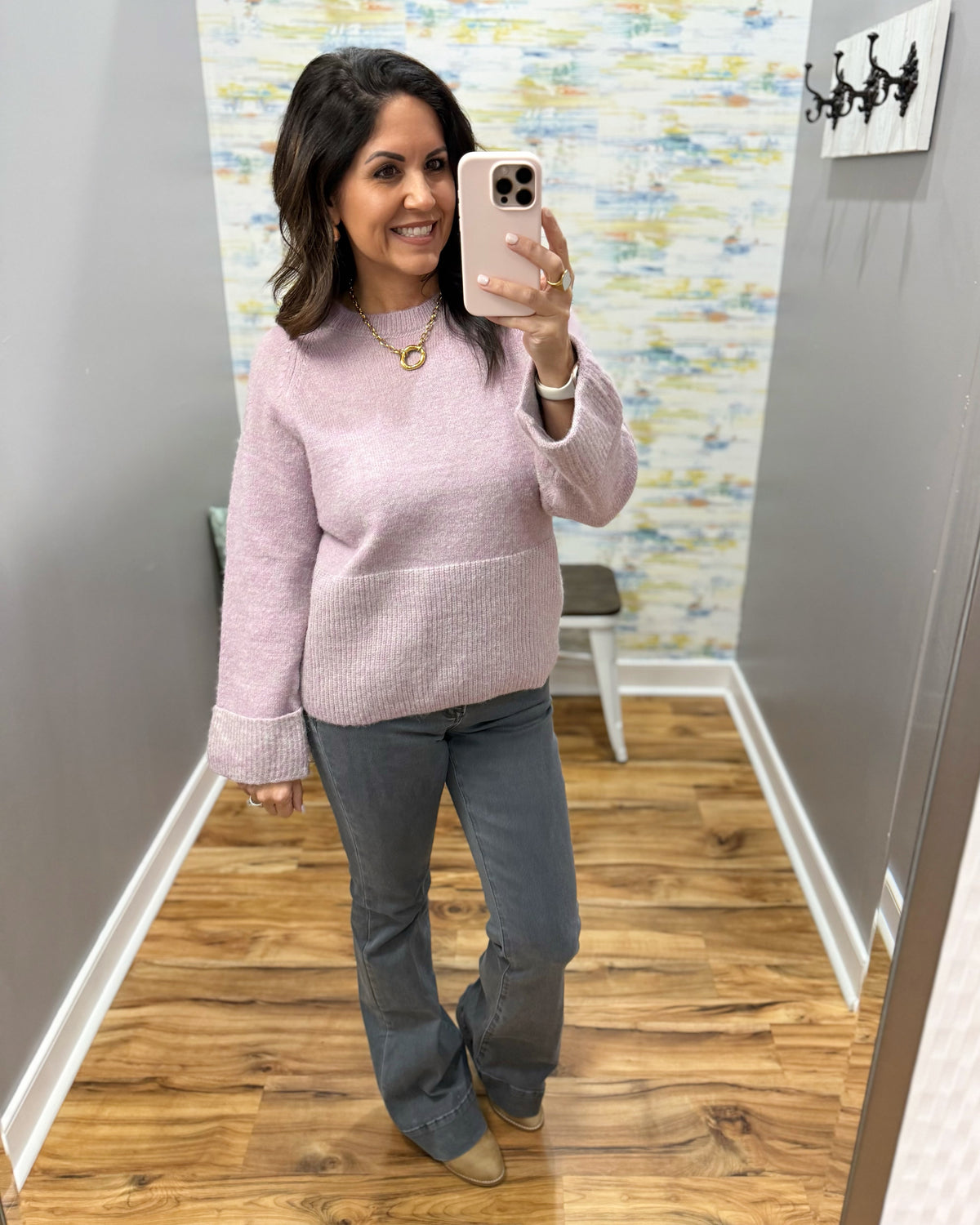A woman takes a mirror selfie wearing a light pink textured knit sweater and gray flared jeans in a fitting room with wood floors and gray walls.