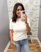 A woman taking a mirror selfie in a fitting room wearing a white short-sleeve crewneck t-shirt and light wash blue jeans.