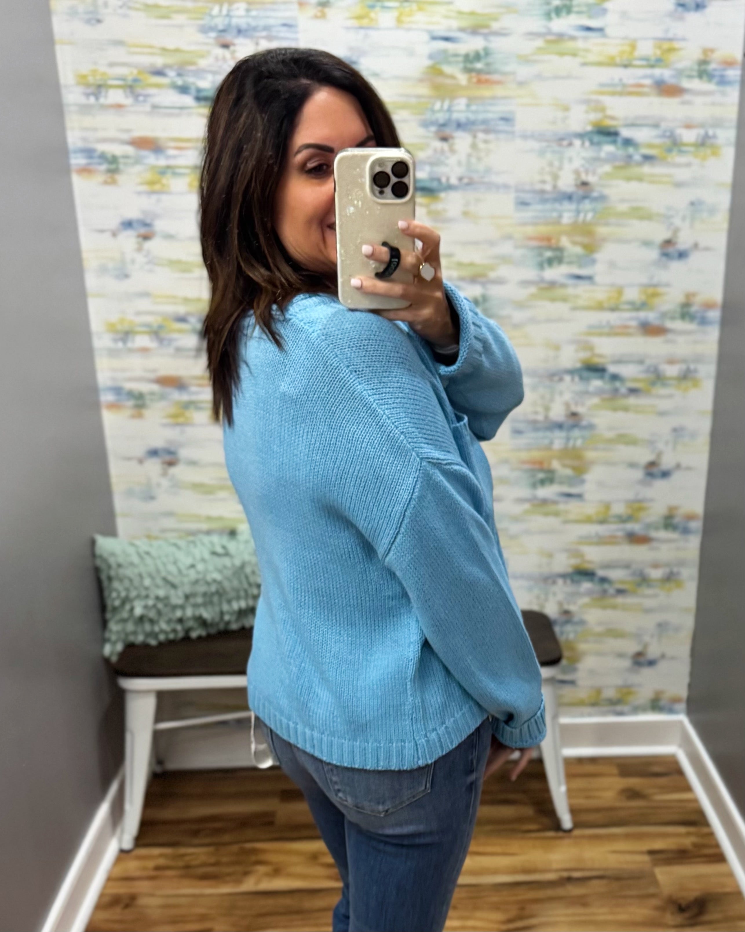 A woman takes a mirror selfie in a fitting room wearing a light blue knit sweater and blue jeans, with a patterned wallpaper background.
