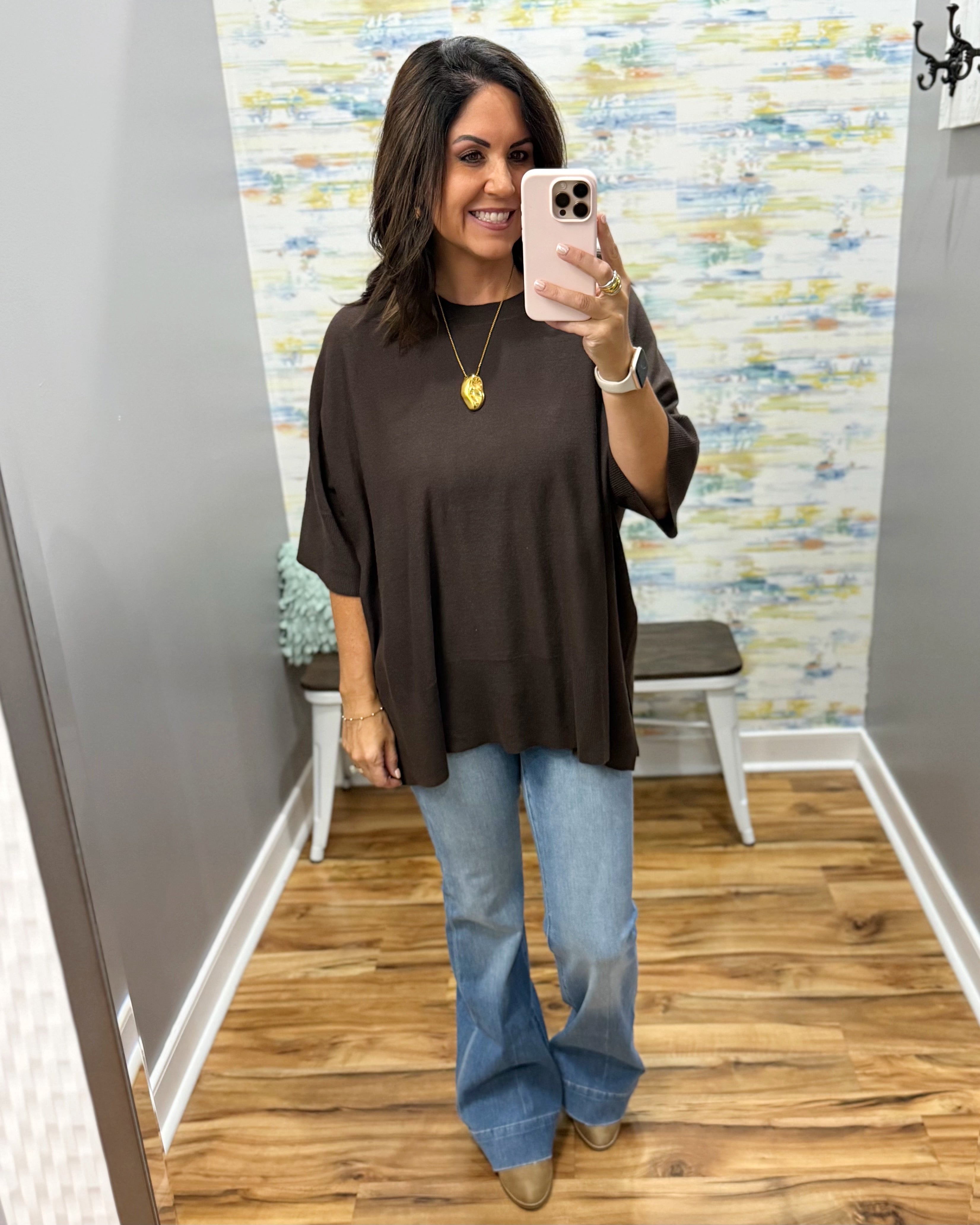 A woman takes a mirror selfie wearing a loose-fitting dark brown short-sleeve top, light-wash blue flare jeans, and tan boots in a fitting room with wood floors and a colorful abstract wallpaper.