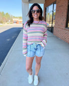 Woman wearing a colorful striped sweater and denim shorts standing on a sidewalk.