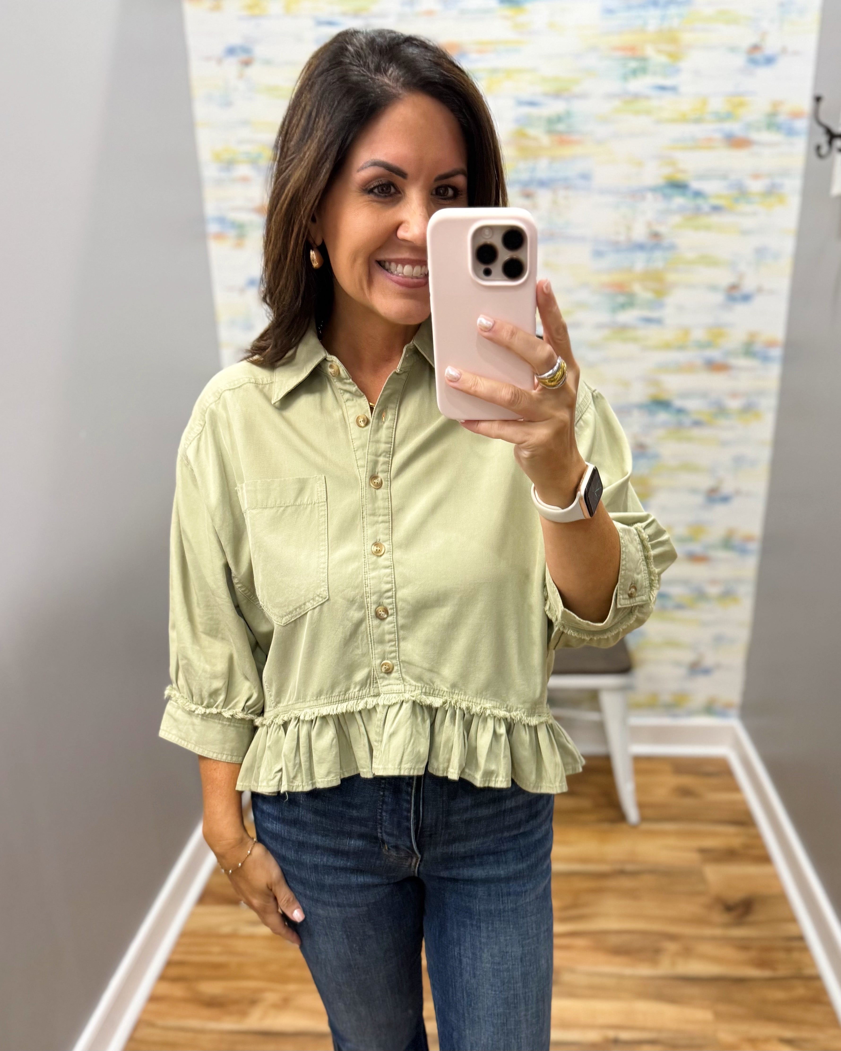 A woman takes a mirror selfie wearing a sage green ruffled blouse with three-quarter sleeves and blue jeans in a fitting room.