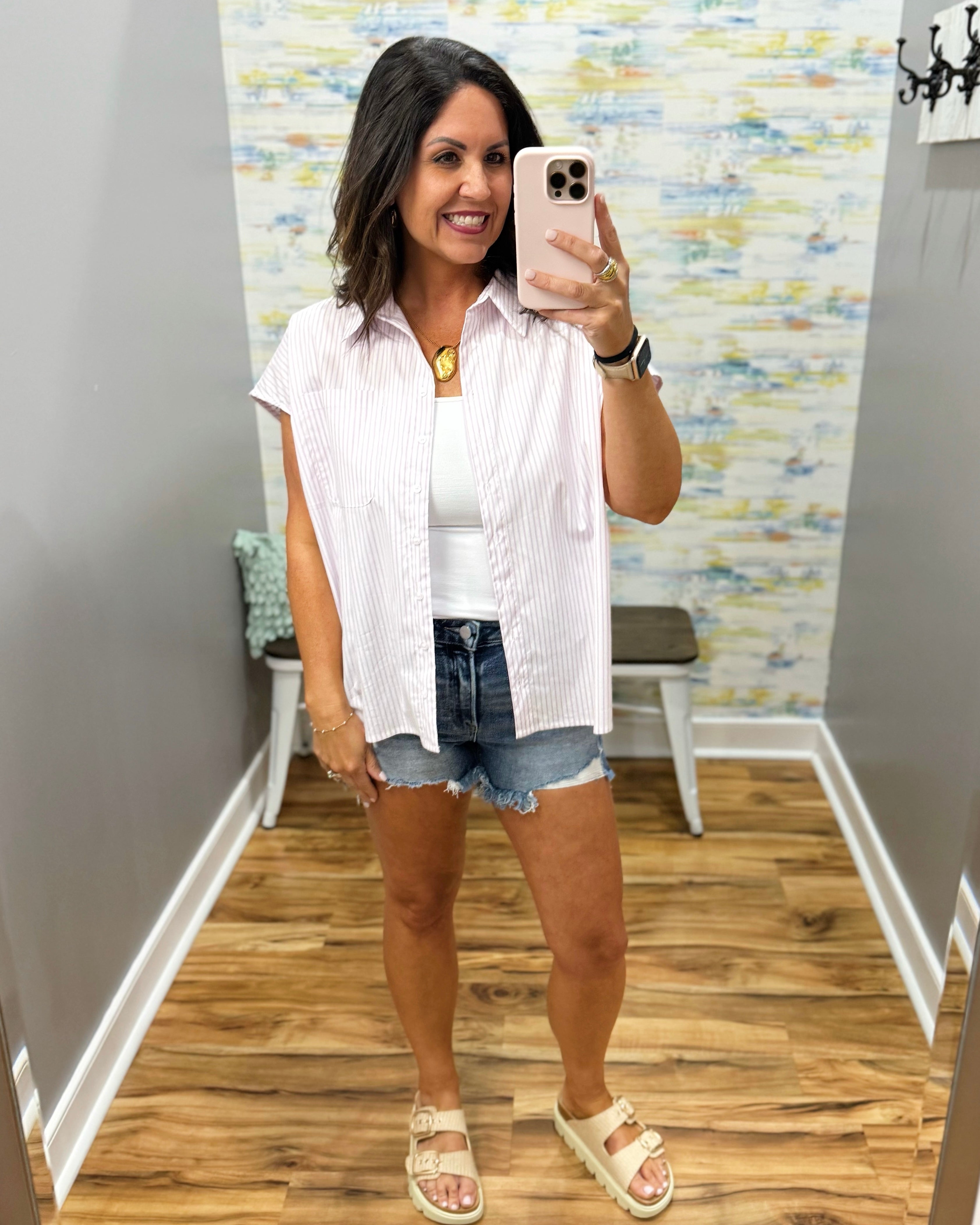 Woman taking a mirror selfie wearing a white button-up shirt, denim shorts, and sandals in a room with a patterned wall.