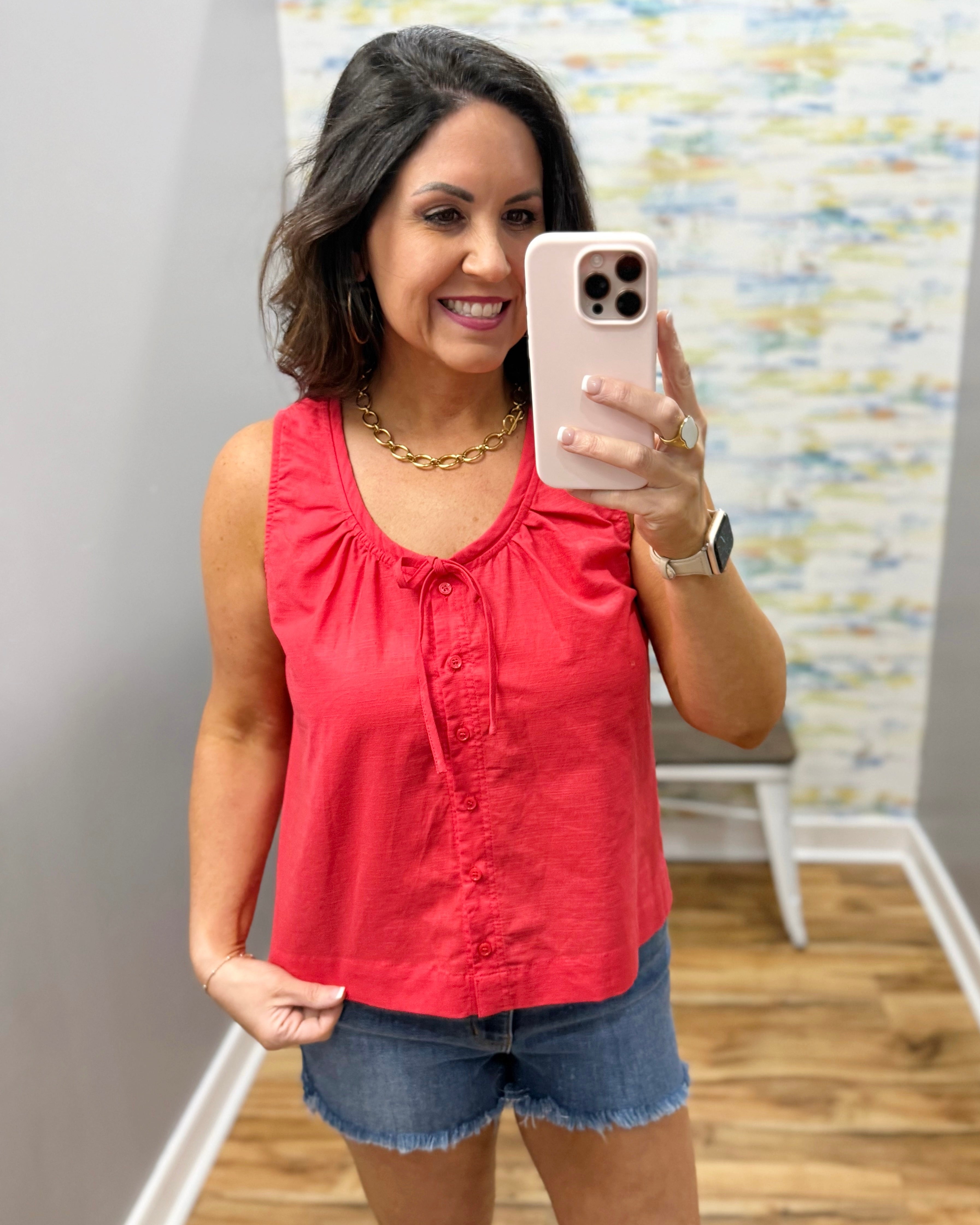 Woman taking a mirror selfie wearing a red sleeveless top and denim shorts.