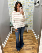 A woman takes a mirror selfie wearing a long-sleeve crewneck sweater with horizontal stripes in cream, light pink, and taupe, paired with blue flare jeans and tan boots.