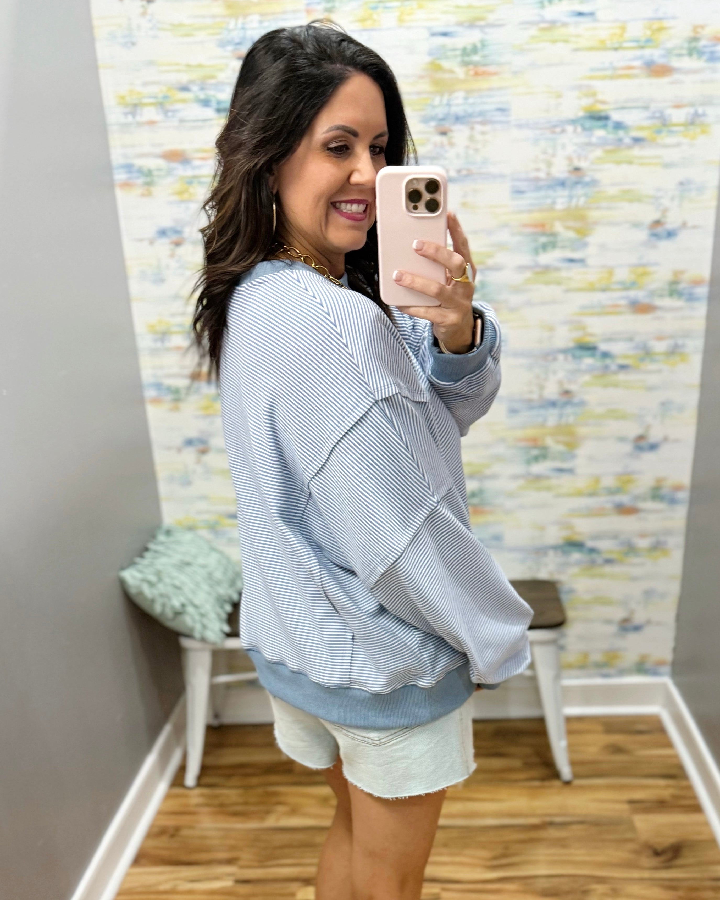 Woman taking a mirror selfie wearing a light blue jacket and white shorts in a room with a textured wall.