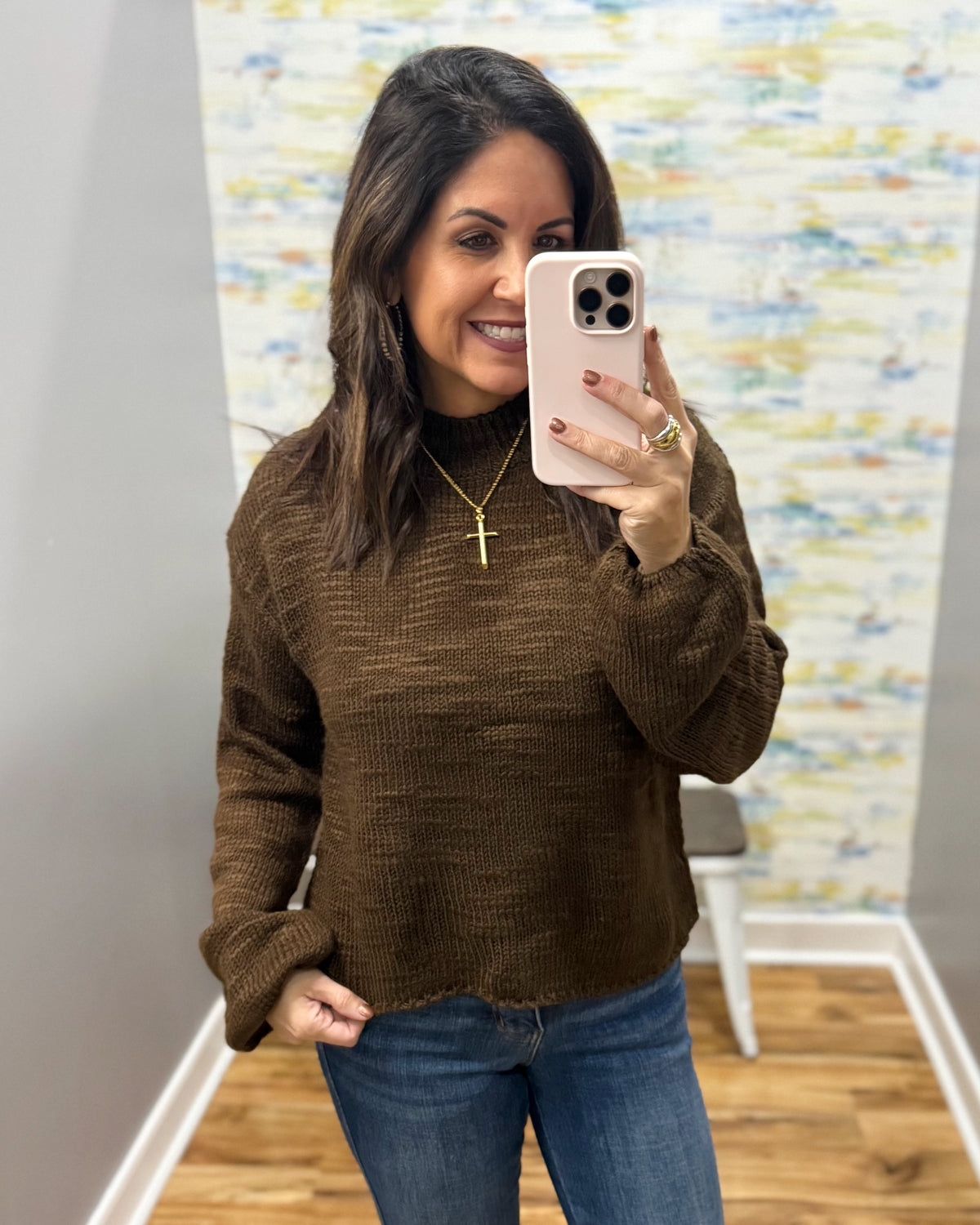 A woman taking a mirror selfie wearing a brown textured knit mock-neck sweater and blue jeans, holding a light pink smartphone.