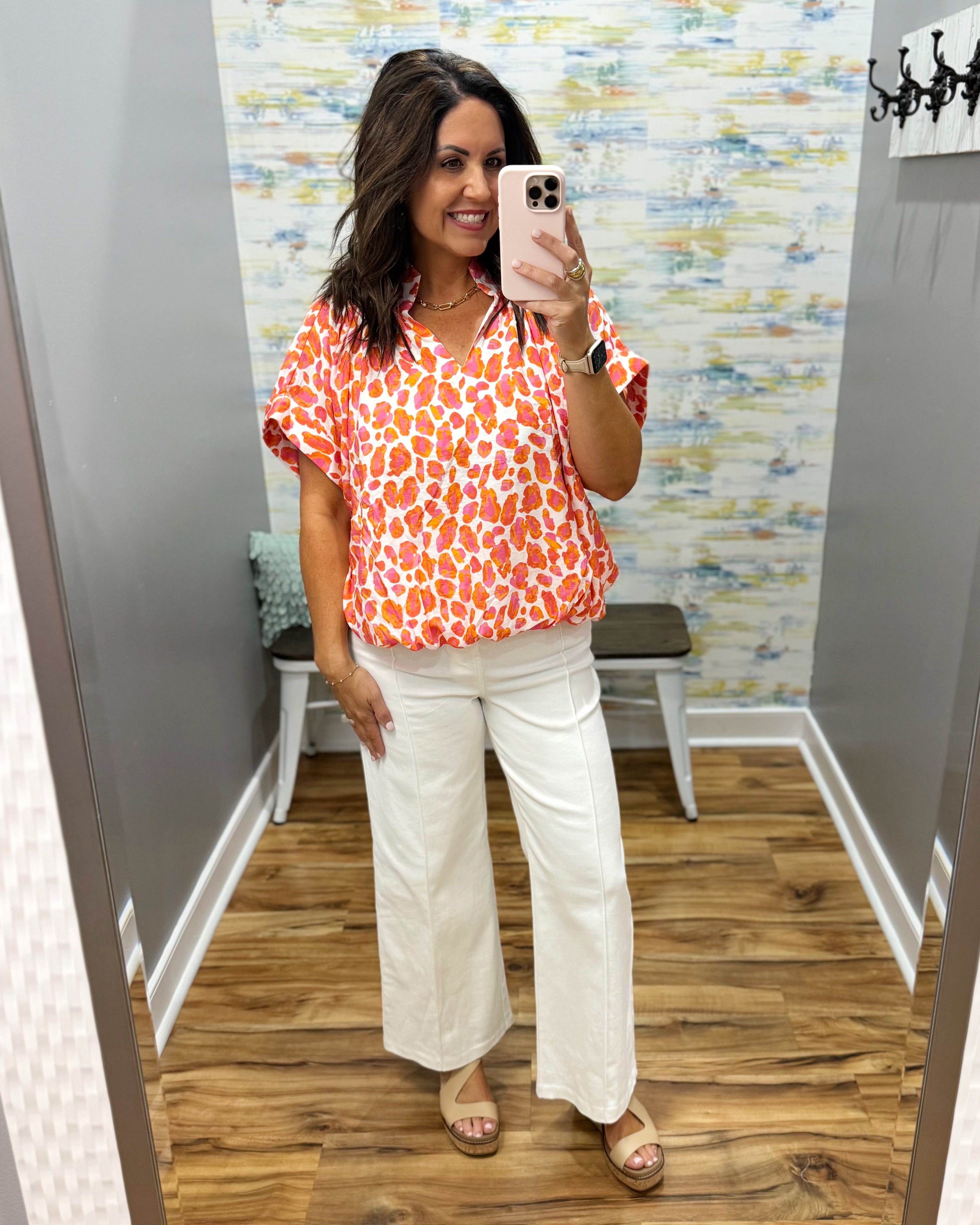 Woman taking a mirror selfie wearing an orange floral blouse and white pants in a room with patterned wallpaper.