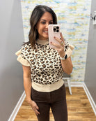 A woman takes a mirror selfie wearing a beige and brown leopard print knit top with ruffled sleeves and dark brown pants in a home setting.