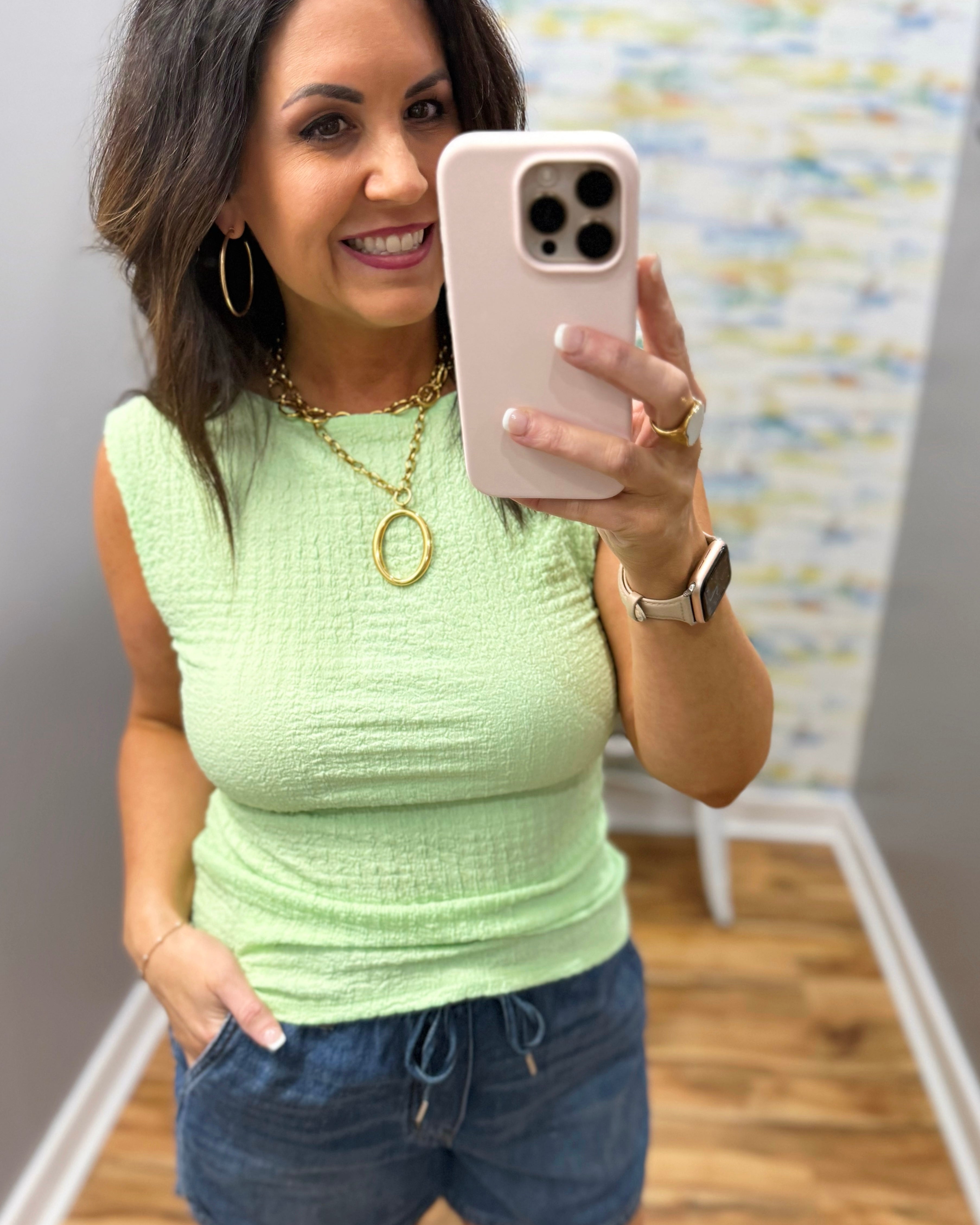 Woman wearing a light green top and blue jeans, holding a pink phone.