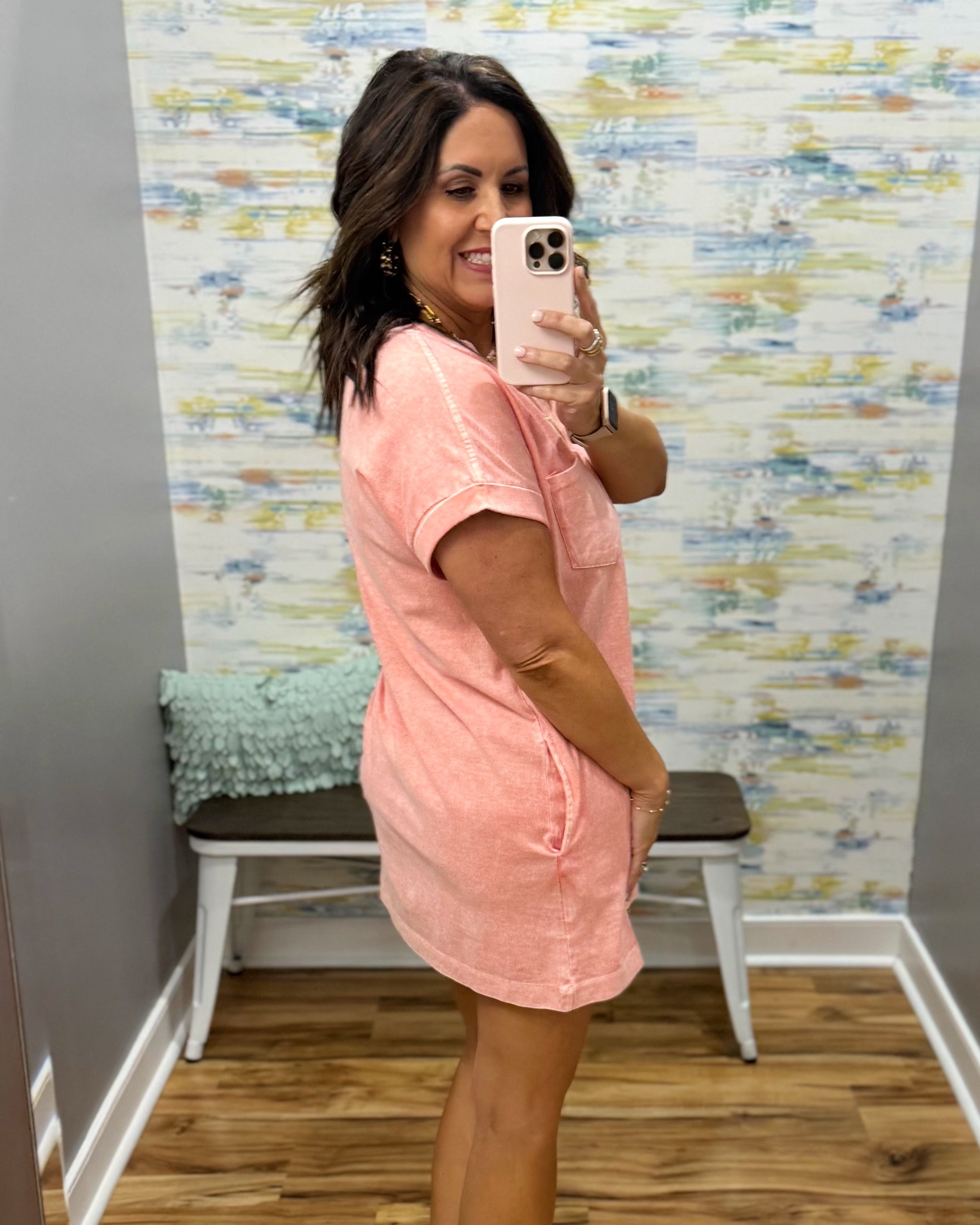 Woman taking a mirror selfie wearing a pink dress in a room with colorful wallpaper.
