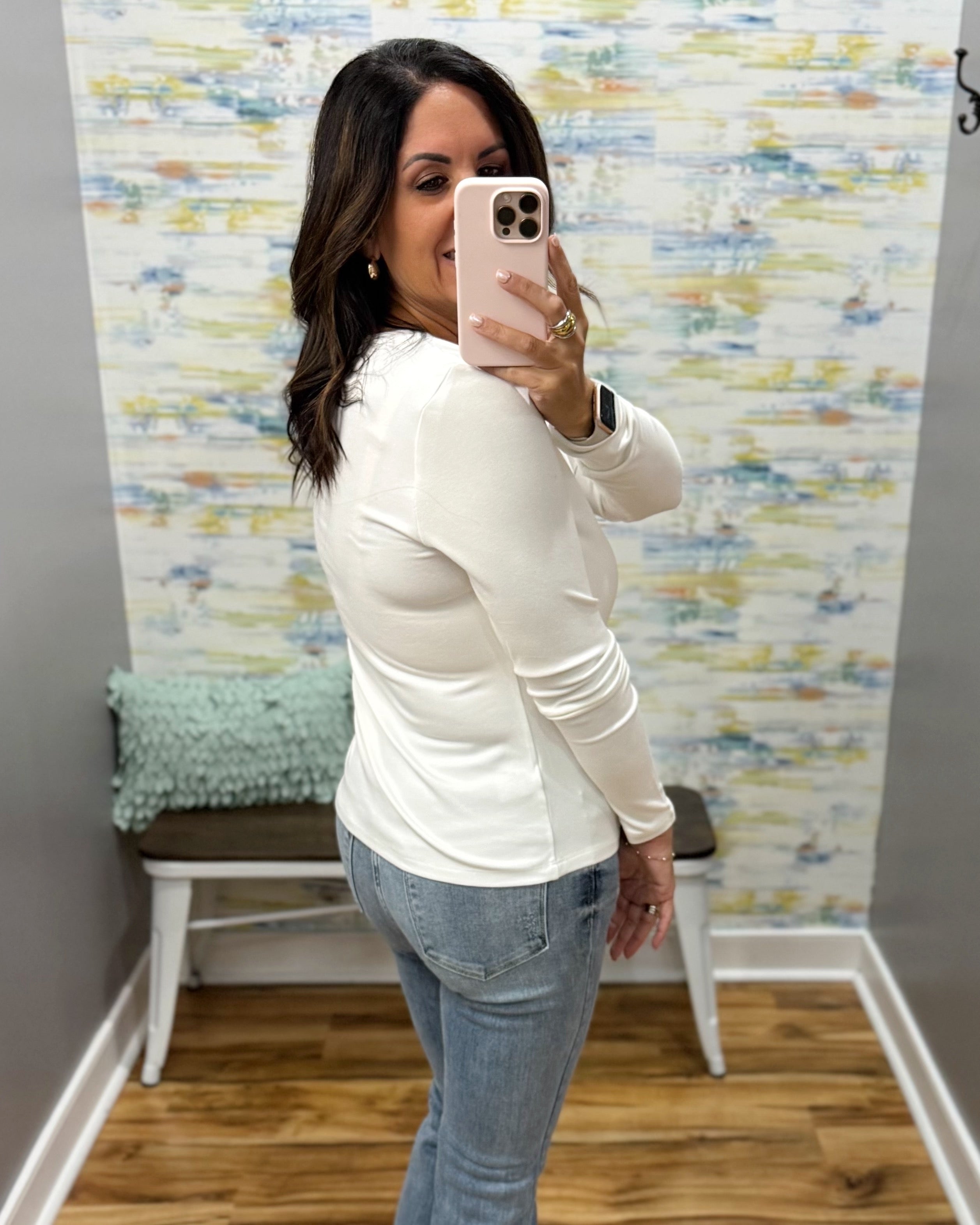 A woman takes a mirror selfie in a fitting room wearing a fitted white long-sleeve top and light blue jeans.
