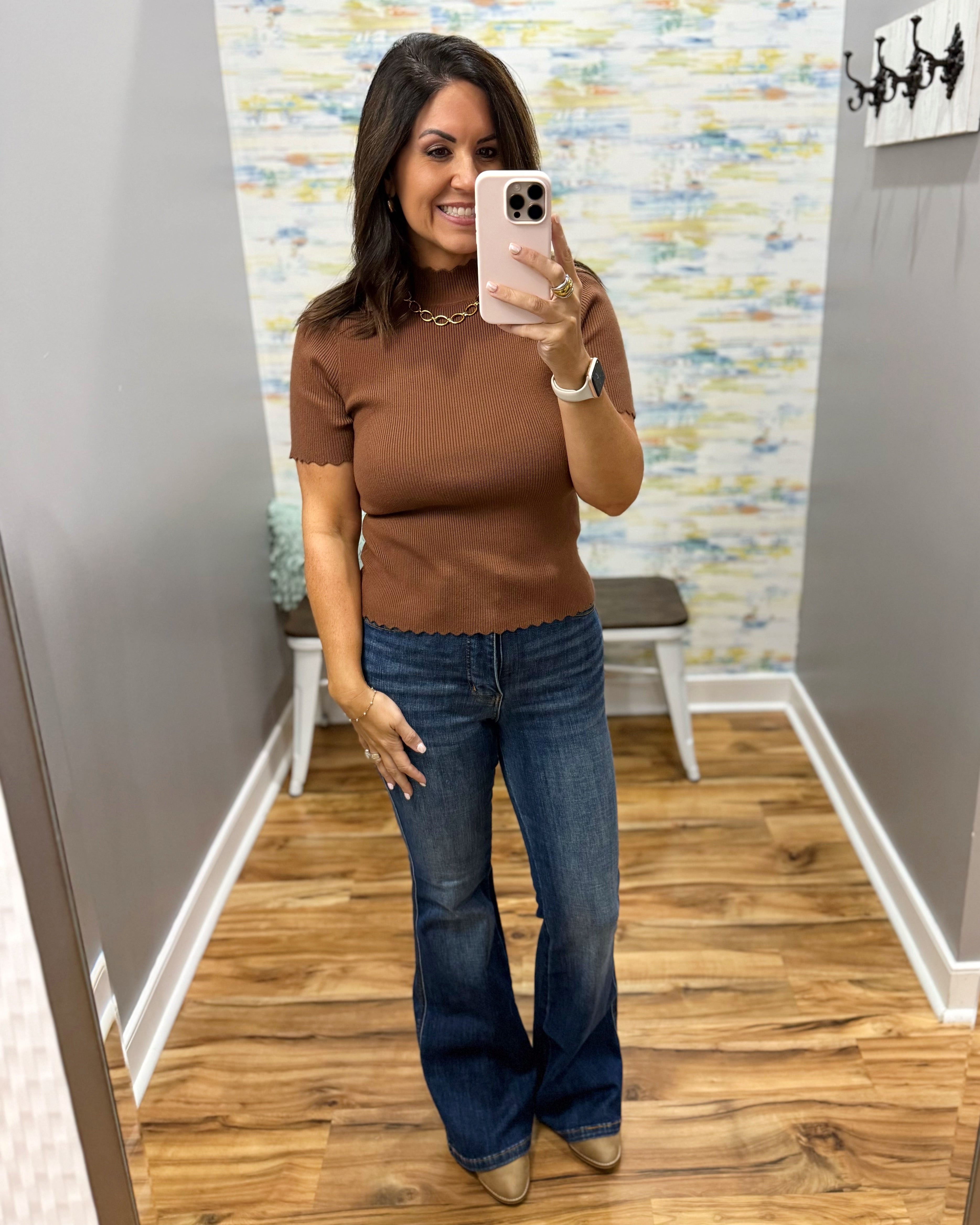 A woman takes a mirror selfie in a fitting room wearing a brown short-sleeve mock neck top and blue flare jeans.