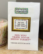 A packaged cross stitch kit labeled 'subversive cross stitch' with the phrase 'did you take your meds today?' shown on the front, photographed on a tabletop next to a candle.